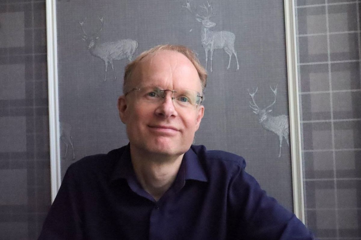 A man Matthew Eagles wearing glasses and a dark shirt in front of a wall with deer illustrations.