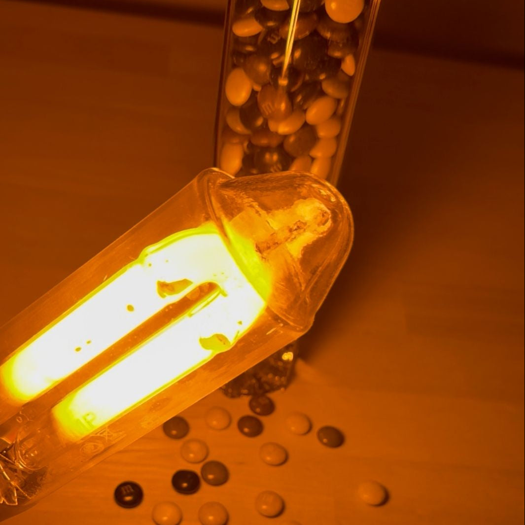A close-up of a glowing fluorescent light bulb in a clear tube, casting a strong warm yellow-orange light onto a wooden surface with scattered round objects and a glass container in the background.